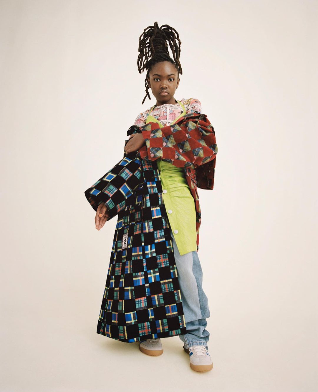 Full-length portrait of a Black female wearing stylish mismatched clothing in a studio. She wears her dreadlocks up in a high bun.