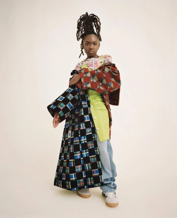 Full-length portrait of a Black female wearing stylish mismatched clothing in a studio. She wears her dreadlocks up in a high bun.