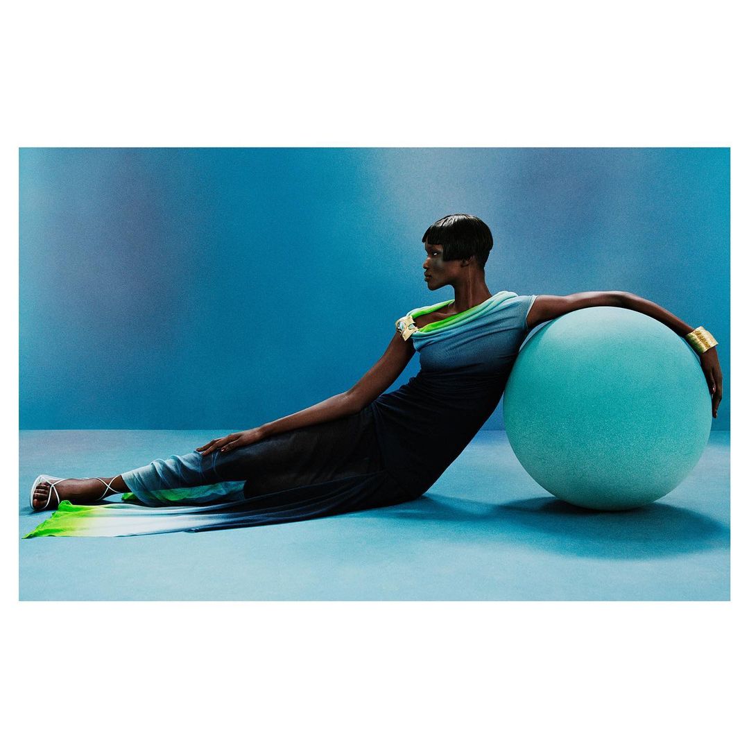 Fashion shot of a Black woman in gradient dress, resting on a large teal sphere in a light blue studio, wearing gold cuff bracelet.