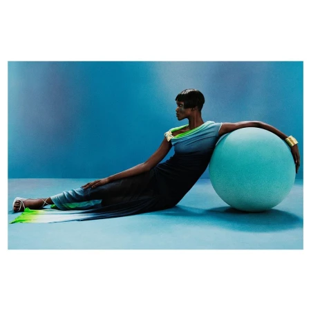 Fashion shot of a Black woman in gradient dress, resting on a large teal sphere in a light blue studio, wearing gold cuff bracelet.