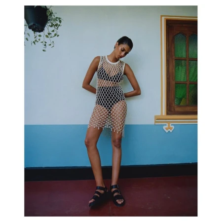 Model wearing a white net dress over black swimwear and sandals standing against a blue and white wall