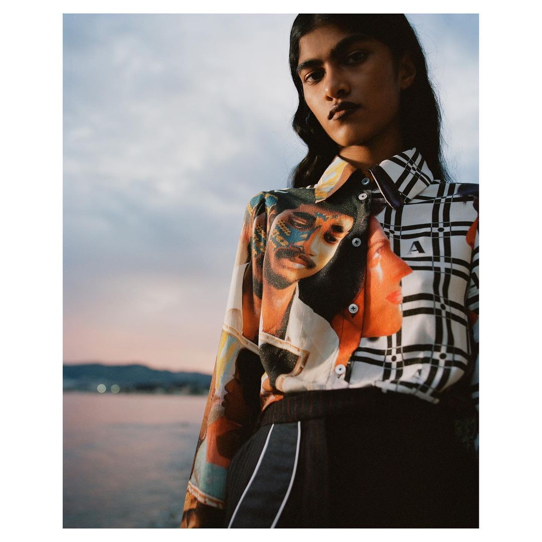 Woman in patterned blouse poses near water at dusk. The blouse has a portrait design on one side and a black-and-white grid pattern on the other.