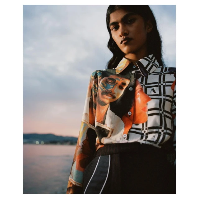 Woman in patterned blouse poses near water at dusk. The blouse has a portrait design on one side and a black-and-white grid pattern on the other.