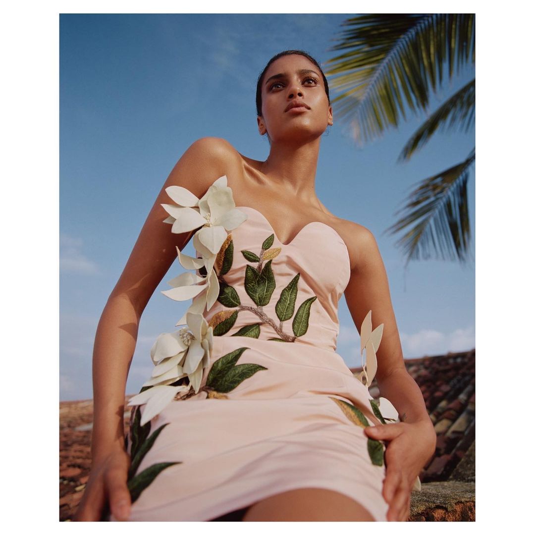 Elegant woman in a floral dress on a rooftop with clear blue skies and palm tree fronds in the background, captured in a low-angle shot.
