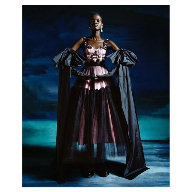Full-length shot of a Black female fashion model in a pink and black tulle dress with statement sleeves, posing against a dark blue backdrop.