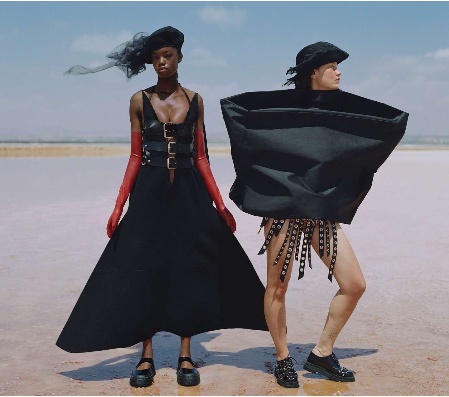 Two fashion models in surreal black outfits pose on a salt flat under a blue sky. One model wears red gloves and platform shoes.