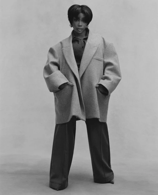 Monochromatic portrait of a Black woman wearing a large grey coat and wide-leg pants against a light grey background.