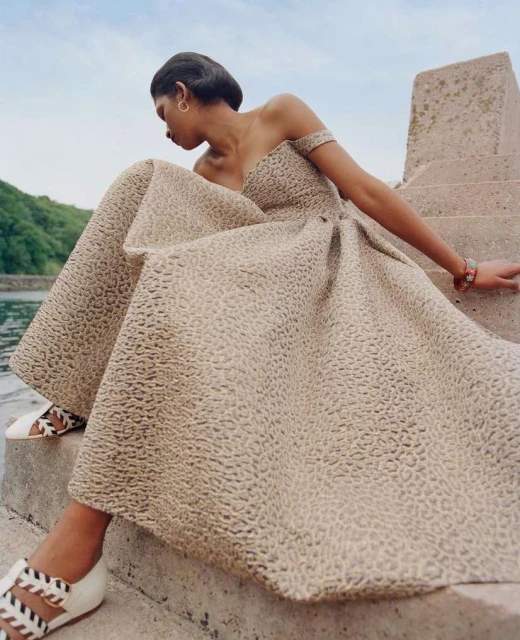 A fashionable woman poses on concrete stairs in a beige dress with textured pattern detail, near water and green landscape in the background.