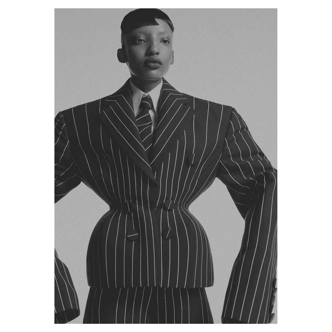Elegant,rogynous person in pinstripe suit with broad shoulders and tailored waist poses in a studio with a neutral gray backdrop.