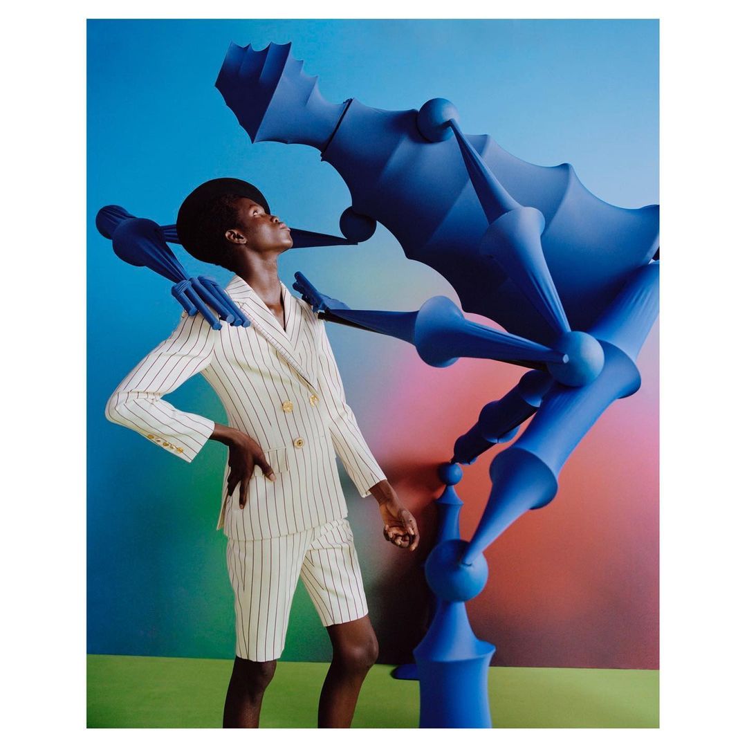 A woman in a pinstripe suit poses in front of a blue architectural sculpture and colorful gradient backdrop in a studio setting.