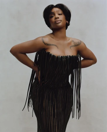 Portrait of SZA in a black fringe dress against a neutral backdrop, showcasing a fashion-forward and elegant studio shot.