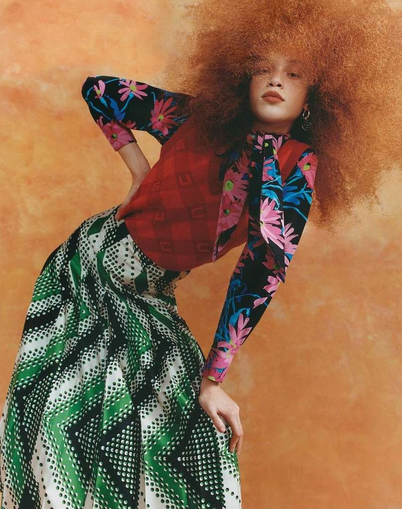 A woman with large curly red hair poses indoors wearing a red, floral print top and a long green skirt against an orange background.