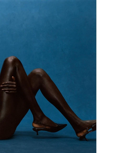 Dark-skinned person poses against a deep blue backdrop, wearing dark, pointed-toe slingback high heels with an artistic heel shape.