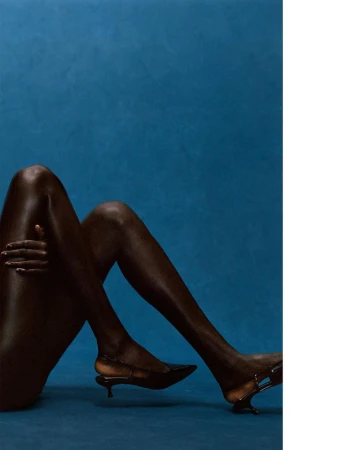 Dark-skinned person poses against a deep blue backdrop, wearing dark, pointed-toe slingback high heels with an artistic heel shape.