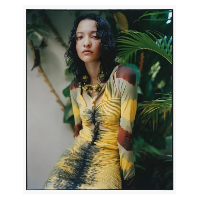 Portrait of an adult woman with dark wavy hair, wearing a yellow dress with long sleeves and a large beaded necklace, posing outdoors among green foliage.