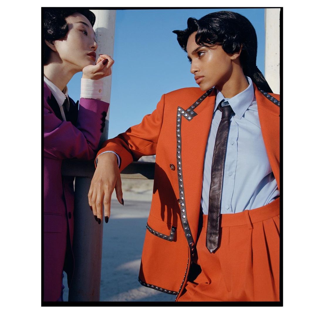 Two people in fashionable attire outdoors. One person wears a bright orange suit with studded trim and a tie. The other wears a purple suit.