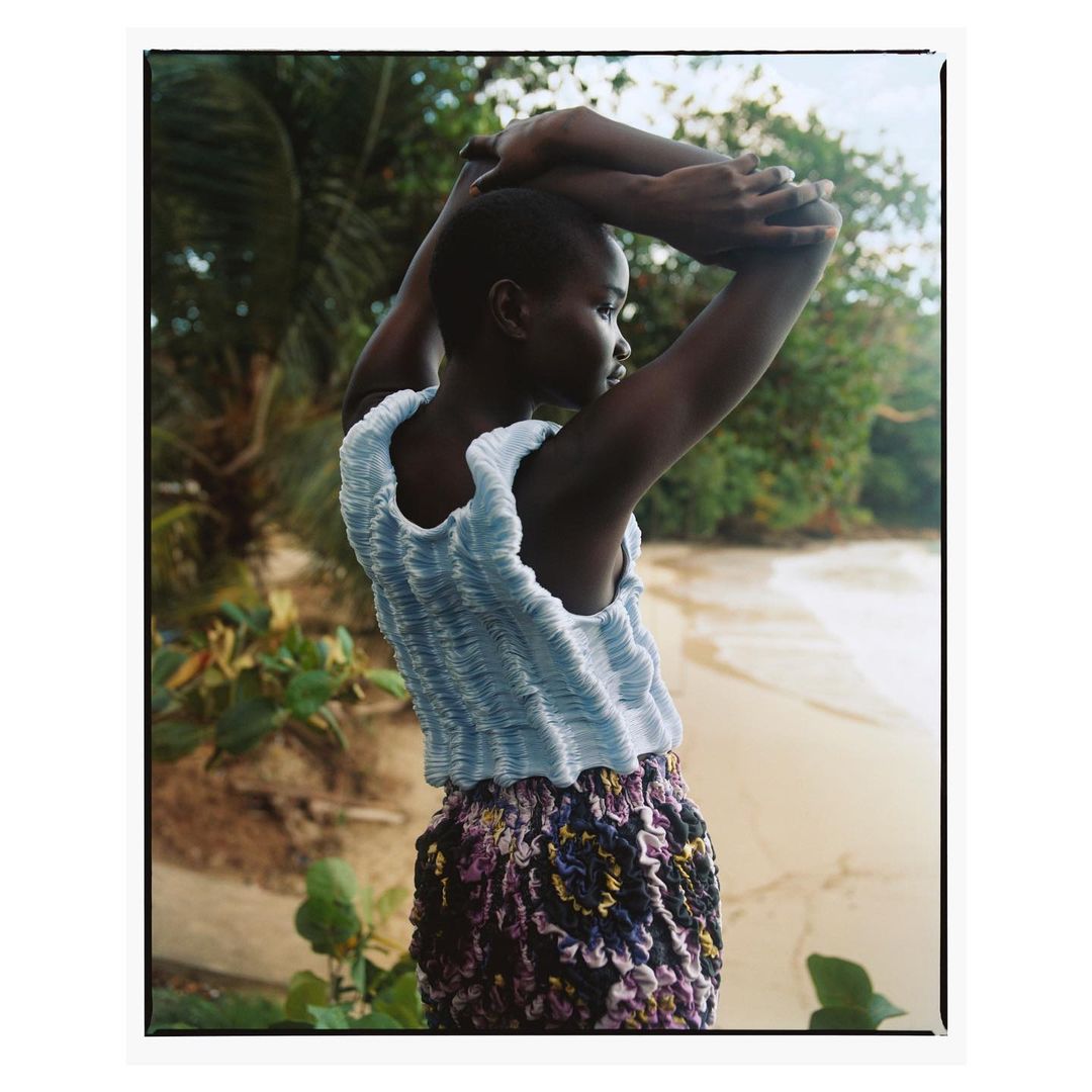 Model with short hair wearing a blue textured top and patterned skirt with arms raised against a lush tropical beach background.