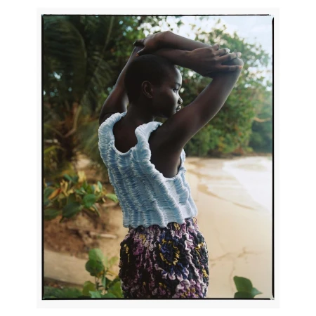 Model with short hair wearing a blue textured top and patterned skirt with arms raised against a lush tropical beach background.