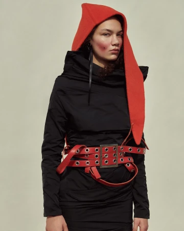 Adult female in a black dress and red hat poses in a studio. The subject is wearing a red belt. The scene is lit by soft lighting.