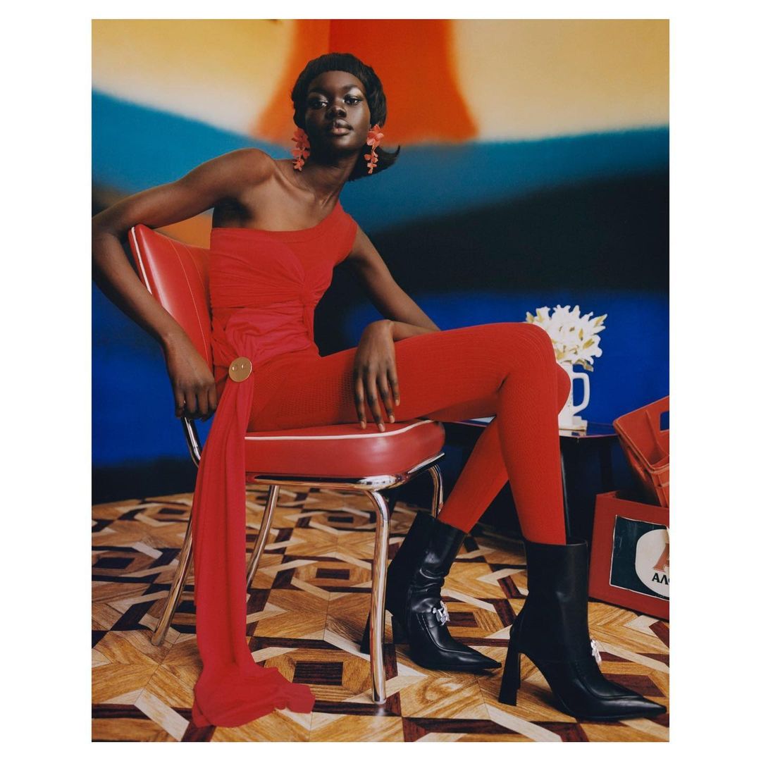 A dark-skinned woman in a red one-shoulder catsuit sits on a red and chrome chair against a blurred studio background in a fashion editorial.