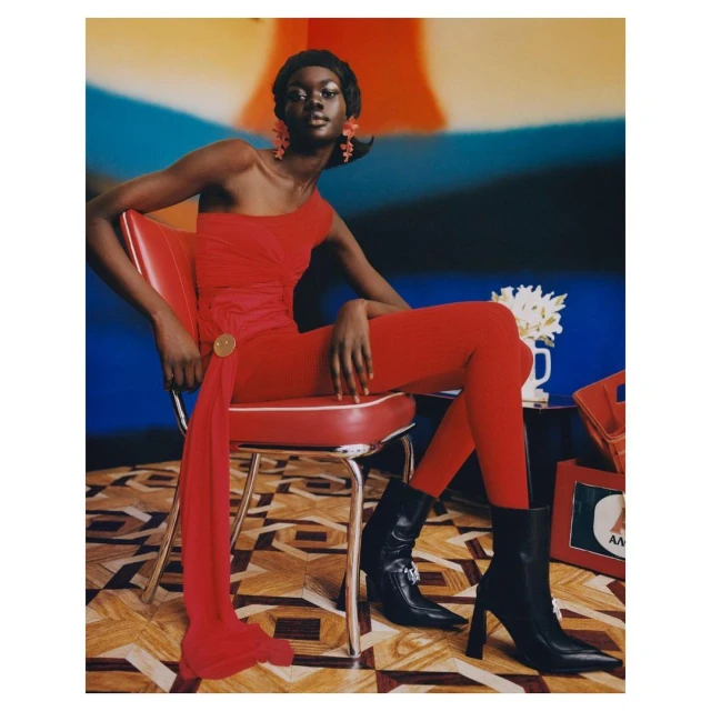 A dark-skinned woman in a red one-shoulder catsuit sits on a red and chrome chair against a blurred studio background in a fashion editorial.