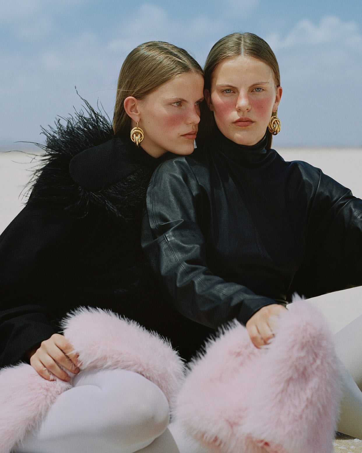 Two women in fashionable black and pink outfits pose together on a bright white surface under a blue sky. They are accessorized with gold earrings.