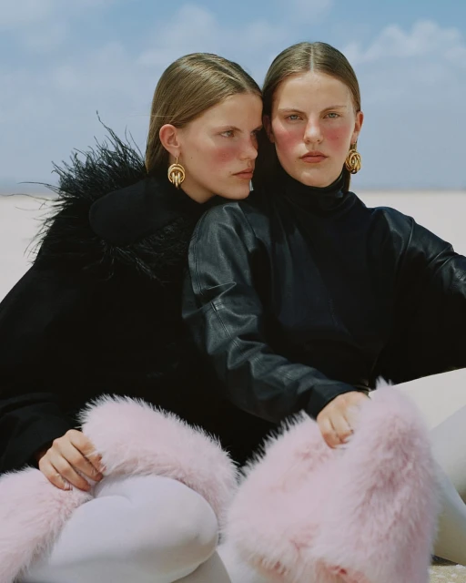 Two women in fashionable black and pink outfits pose together on a bright white surface under a blue sky. They are accessorized with gold earrings.