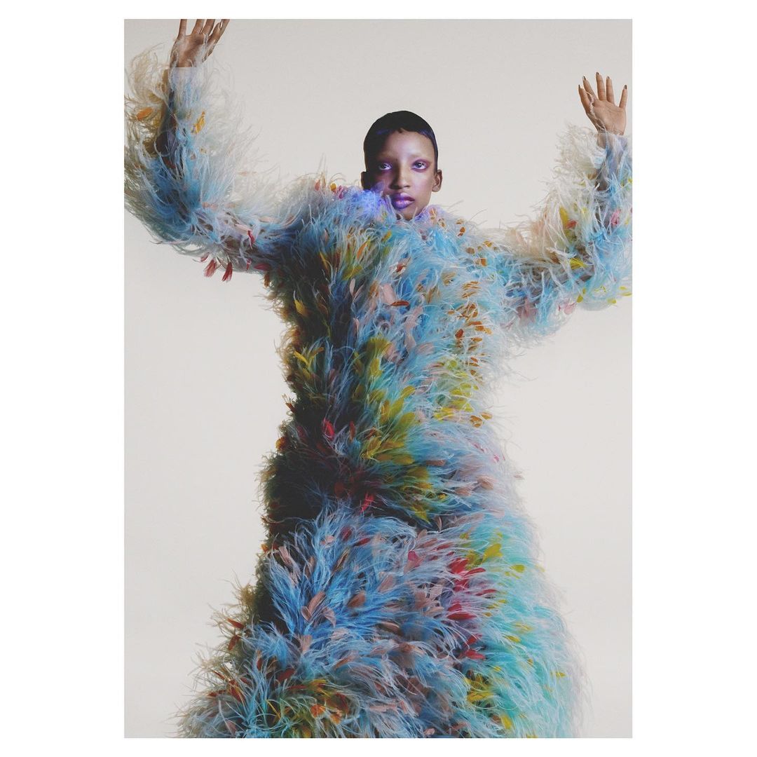 Woman in a feathered blue and yellow outfit poses against a neutral backdrop with her arms raised in a fashion or artful display.