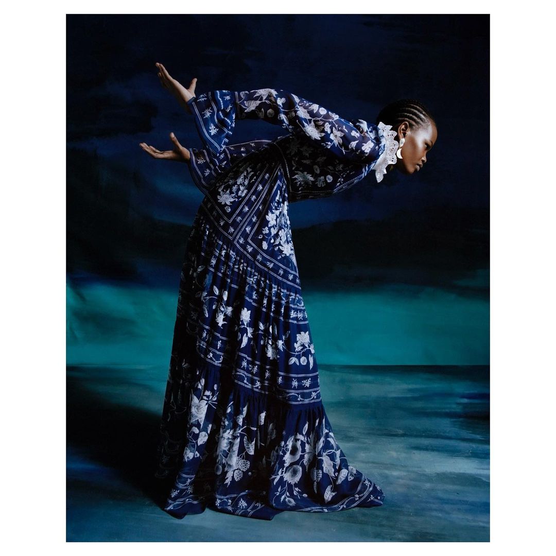 An adult female poses with arms raised wearing a long blue-patterned dress against a blue-green backdrop in a fashion editorial shot.