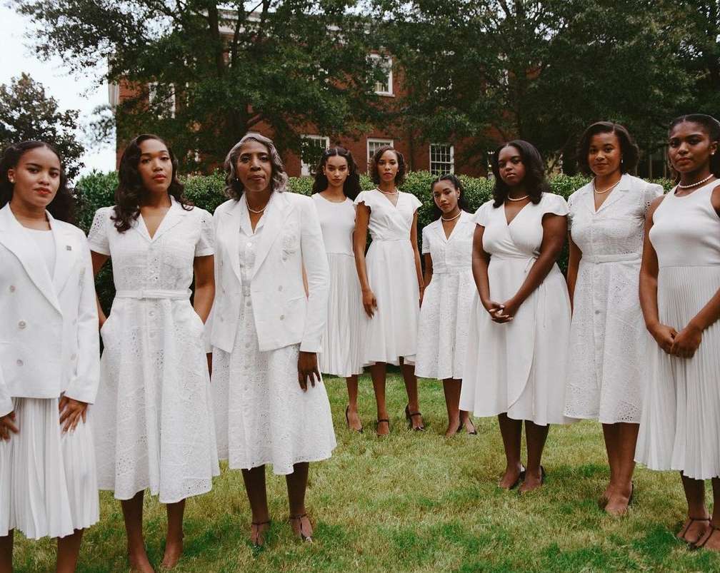 Eight women stand in an outdoor setting. They are wearing white dresses and posed together with a brick building in the background.