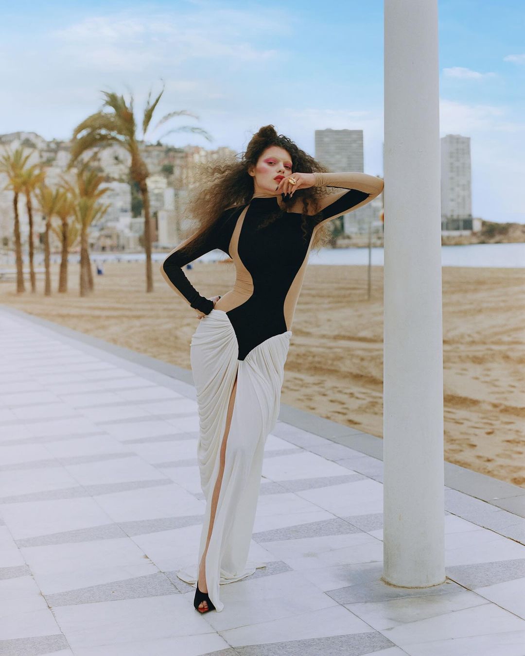 Fashion photography of a woman in a black and white dress posing against a white pillar, with palm trees and city buildings in background.