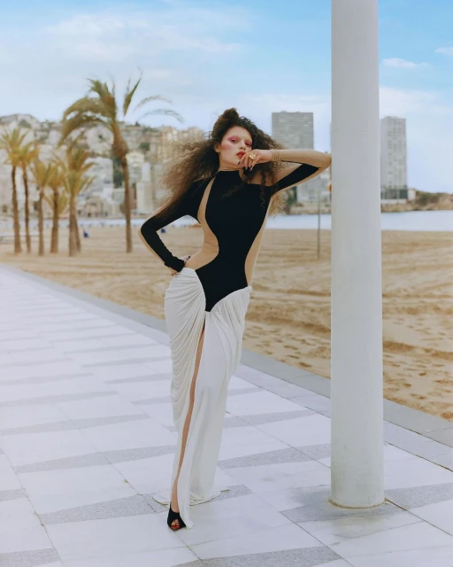 Fashion photography of a woman in a black and white dress posing against a white pillar, with palm trees and city buildings in background.