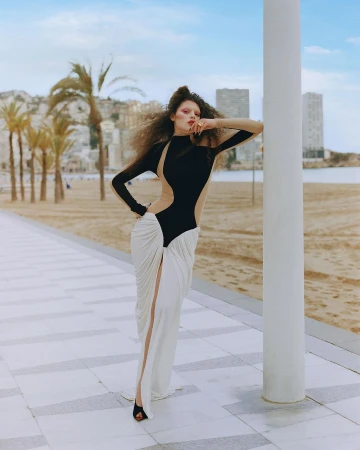 Fashion photography of a woman in a black and white dress posing against a white pillar, with palm trees and city buildings in background.