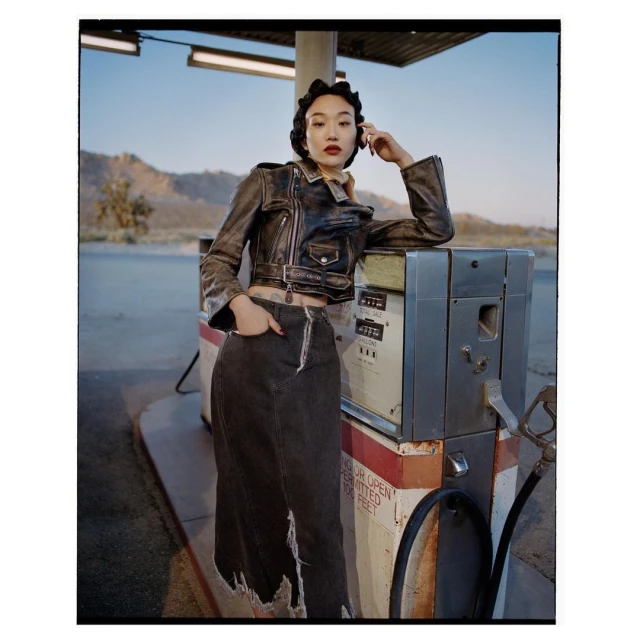 A young Asian woman poses near an old-fashioned gas pump wearing a leather jacket and long denim skirt against a desert landscape.