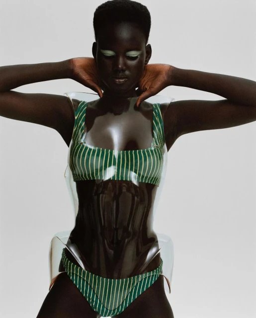 Fashion shot of a Black woman wearing a green and white striped bikini with transparent plastic details against a neutral background.