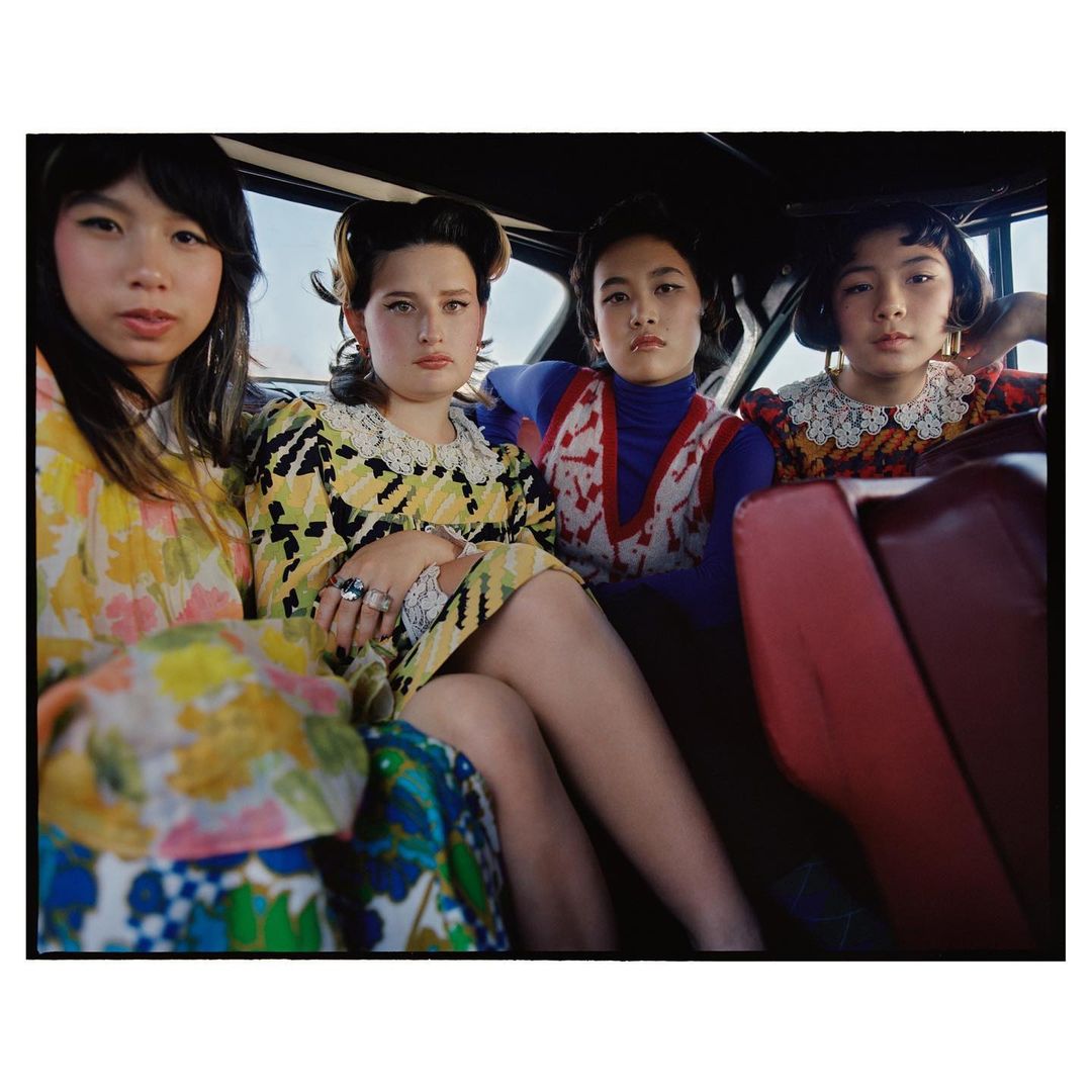 Four young women pose inside a car with retro fashion, yellow, blue, and red floral print dresses, and vintage hairstyles in a photo studio.