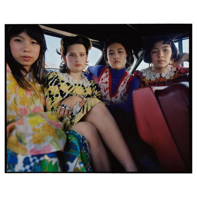 Four young women pose inside a car with retro fashion, yellow, blue, and red floral print dresses, and vintage hairstyles in a photo studio.