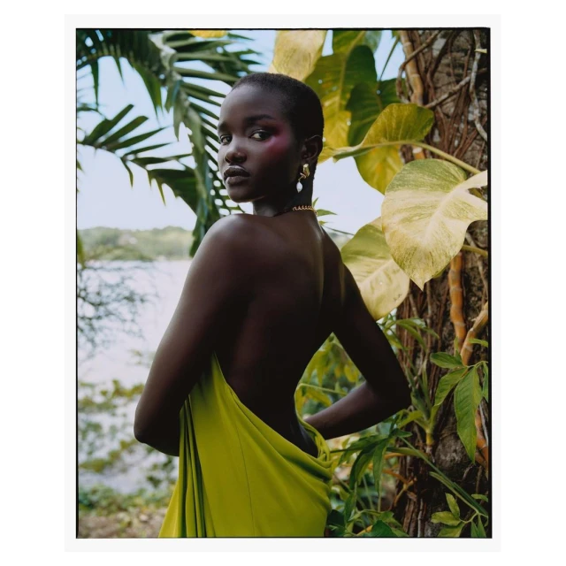 Elegant portrait of a dark-skinned woman with pink makeup and bare shoulders, draped in vibrant yellow fabric, amidst tropical foliage and greenery.
