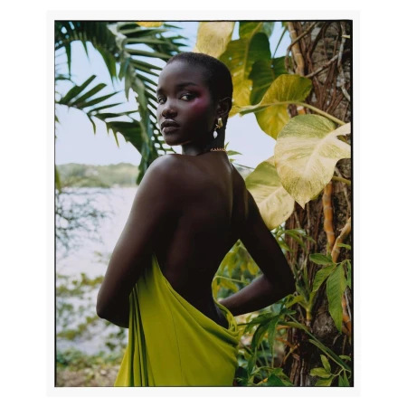 Elegant portrait of a dark-skinned woman with pink makeup and bare shoulders, draped in vibrant yellow fabric, amidst tropical foliage and greenery.