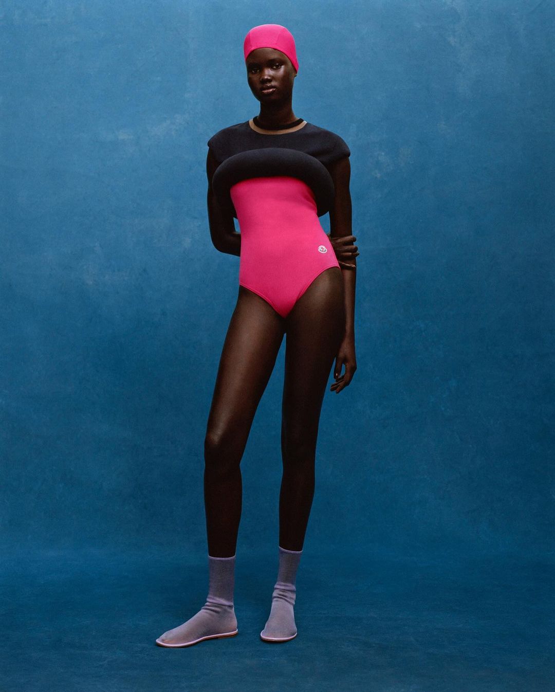 A full-length portrait showcases a Black female model in pink swimwear, paired with lavender socks, against a textured blue backdrop.