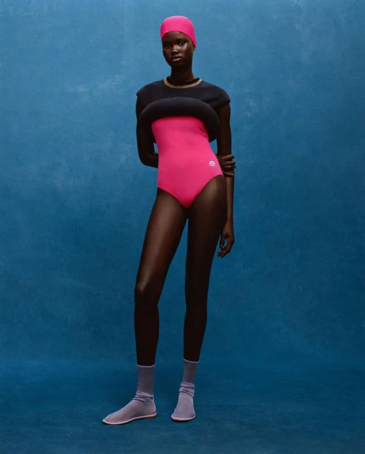 A full-length portrait showcases a Black female model in pink swimwear, paired with lavender socks, against a textured blue backdrop.
