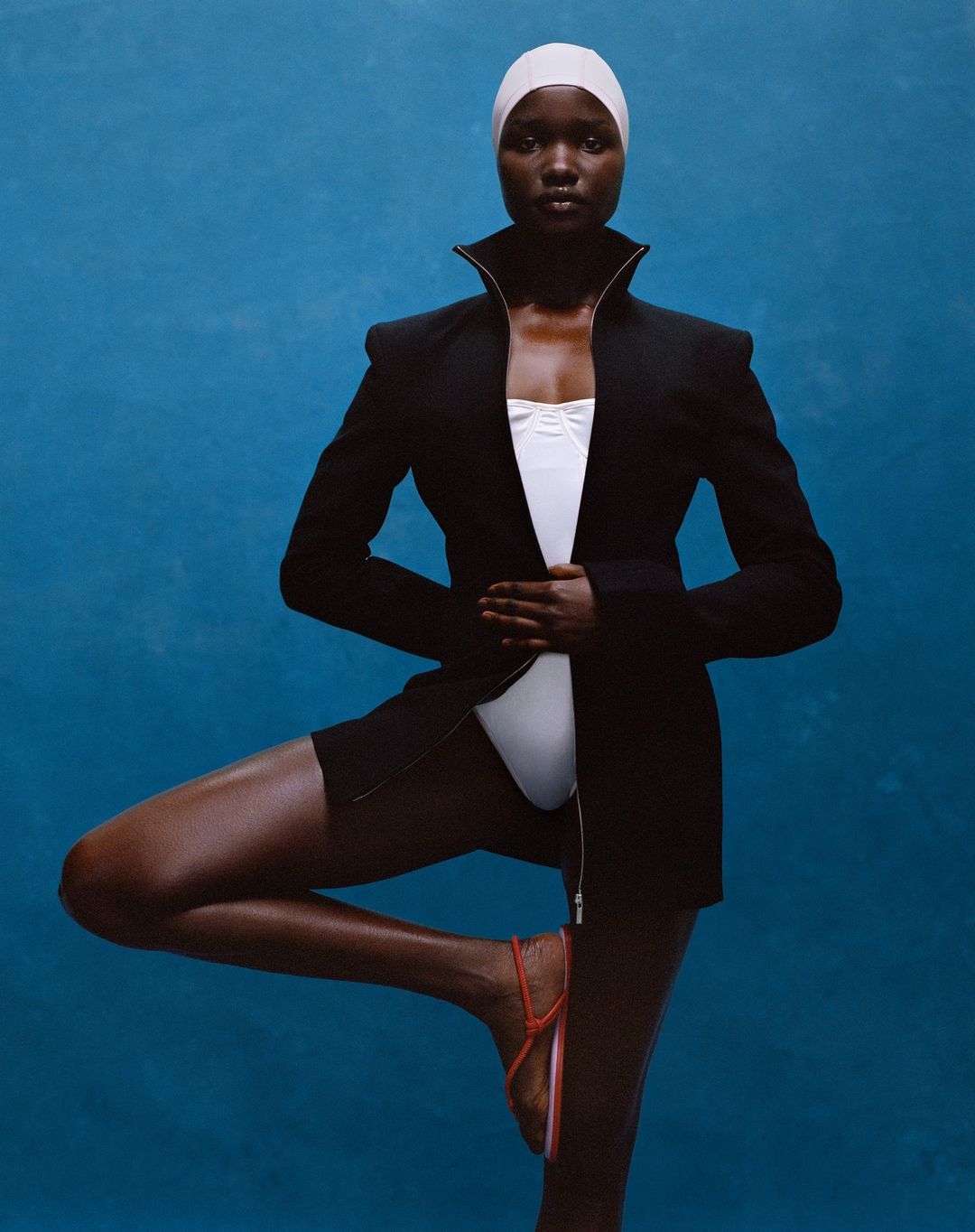 Woman balances in a black blazer, white swimsuit, and swim cap against a blue background in a studio portrait, exhibiting swimwear fashion.