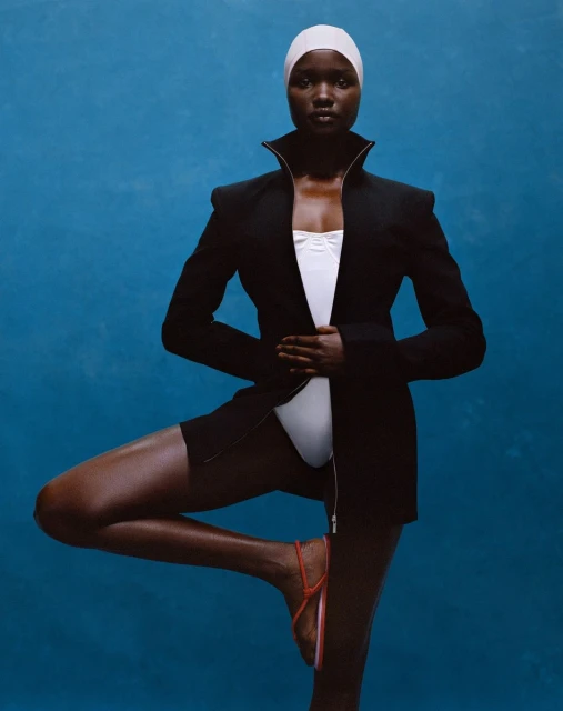 Woman balances in a black blazer, white swimsuit, and swim cap against a blue background in a studio portrait, exhibiting swimwear fashion.