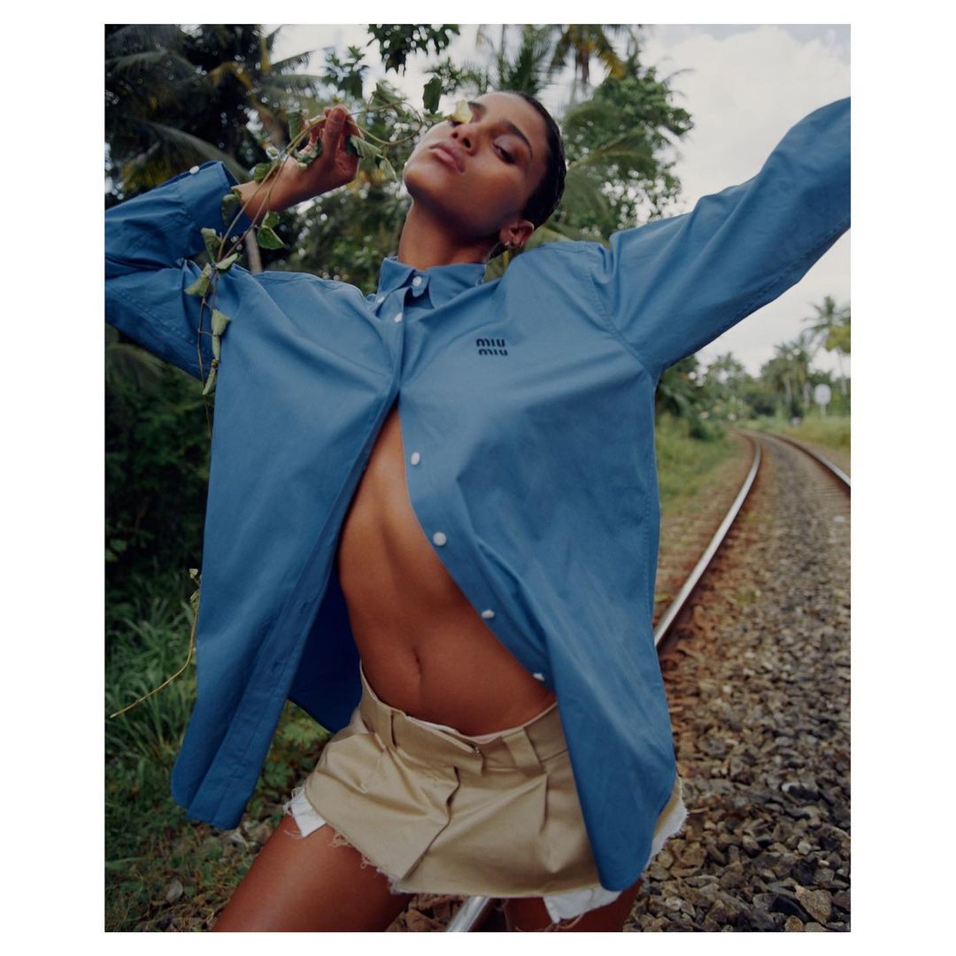 A tanned adult female poses on railroad tracks in an unbuttoned blue shirt and khaki shorts with green vegetation in the background.