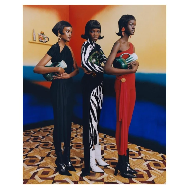 Three black female models in patterned clothing pose indoors holding jugs. The scene features vibrant color blocking and retro styling.