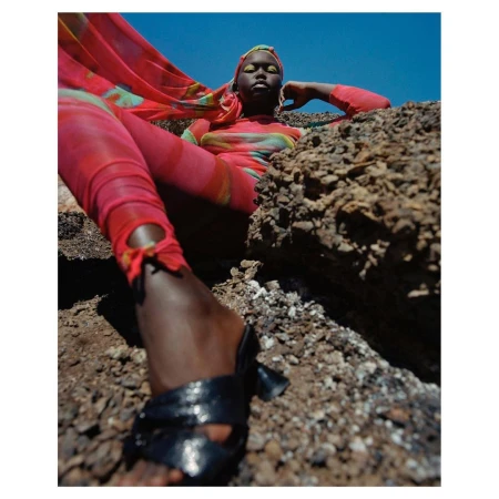 Model in vibrant pink patterned apparel and headscarf reclining on rocky terrain against a clear blue sky, photographed by Nadine Ijewere.