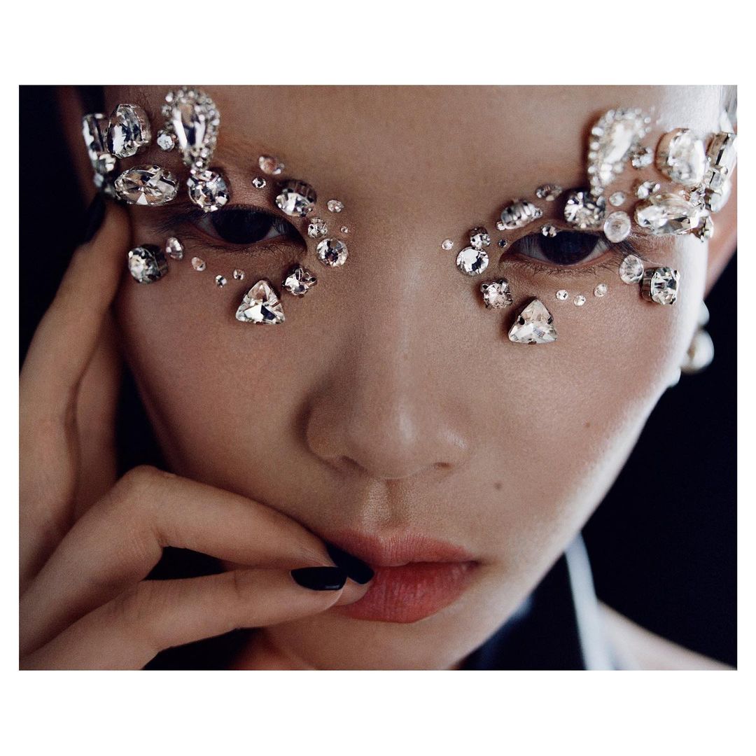 Close-up of a woman's face adorned with sparkling rhinestones around the eyes, fingers near her lips, and a black background.