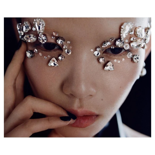 Close-up of a woman's face adorned with sparkling rhinestones around the eyes, fingers near her lips, and a black background.