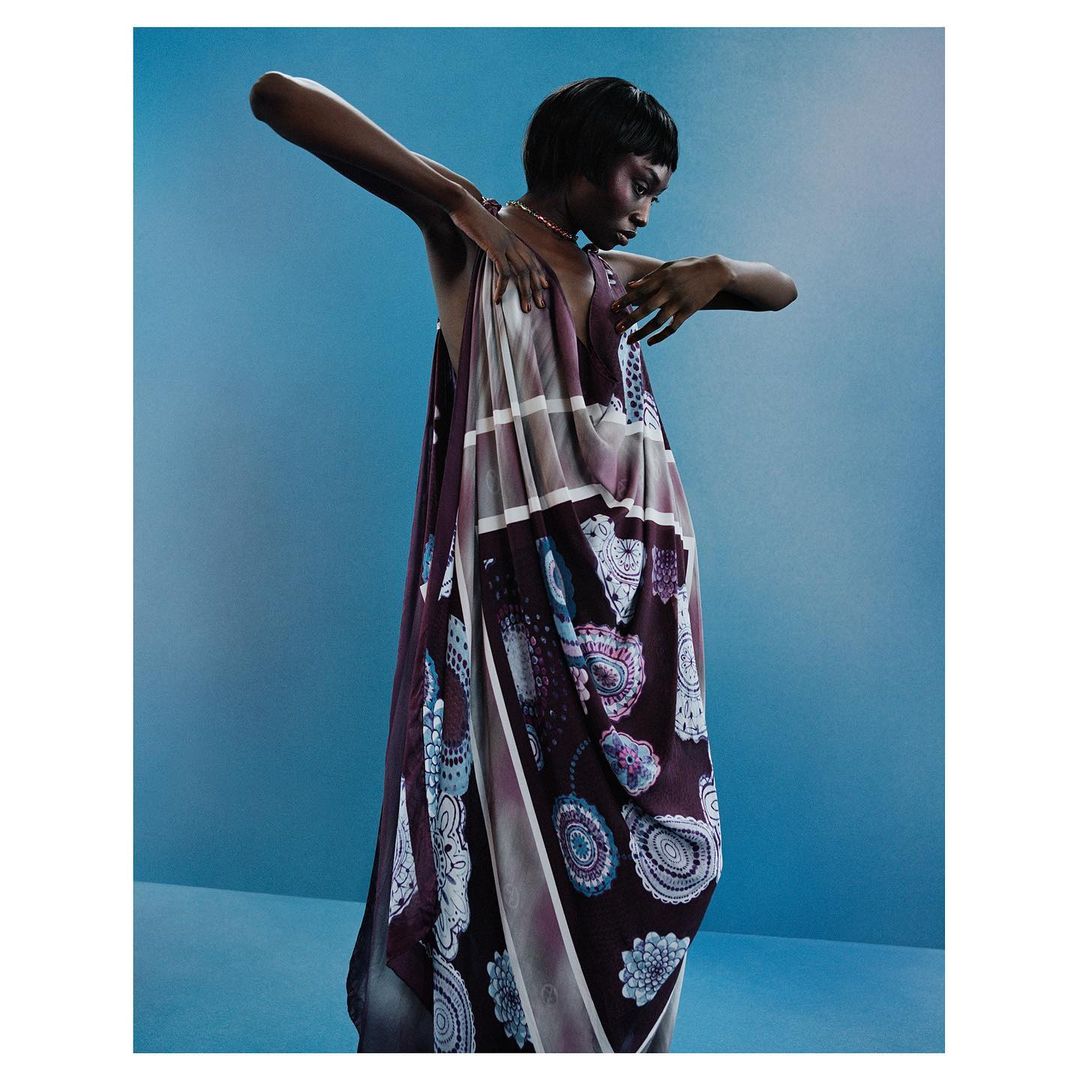 Black woman wearing a patterned draped dress posing against a solid blue studio backdrop in a portrait by Nadine Ijewere