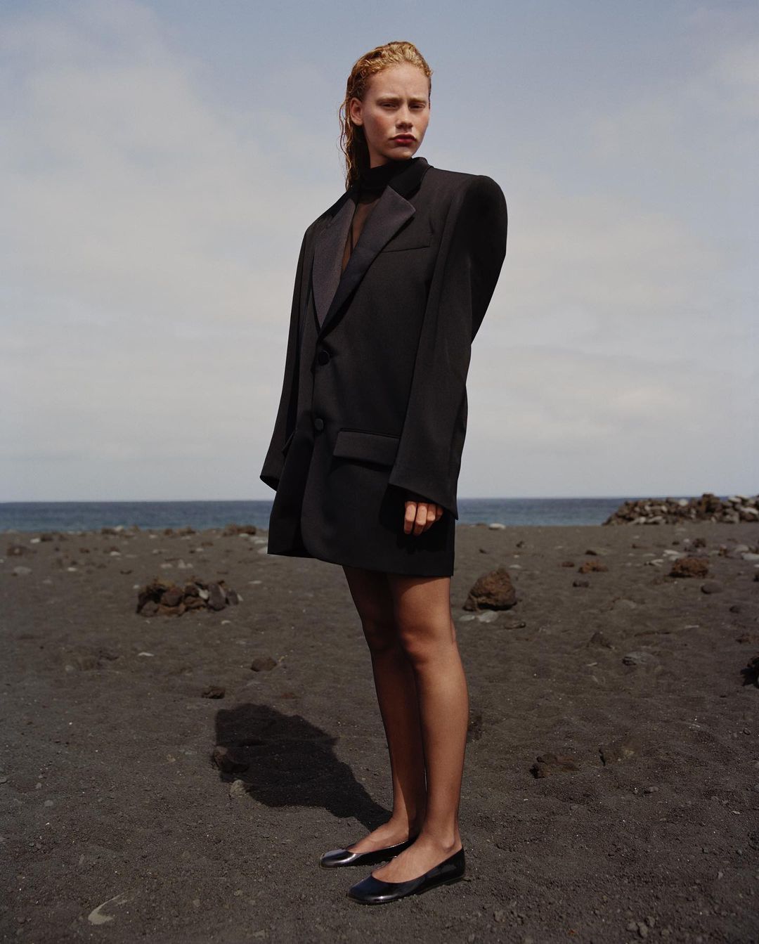 A fair-skinned woman in an oversized black blazer and black flats stands on a dark sand beach under a cloudy blue sky by the ocean.